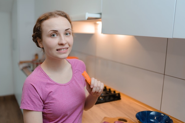 Chica pelirroja hermosa comiendo zanahorias en la cocina. Estilo de vida saludable