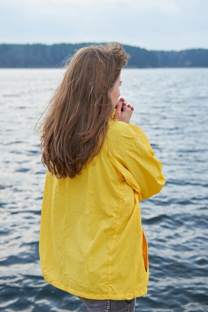 Chica pelirroja en impermeable amarillo calentando las manos, de pie cerca del lago en el desagradable clima frío de otoño