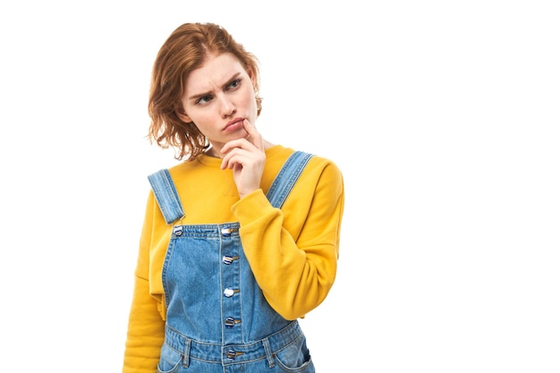 Una chica pelirroja inteligente con barbilla casual piensa que las dudas toman una decisión aislada en el fondo del estudio blanco