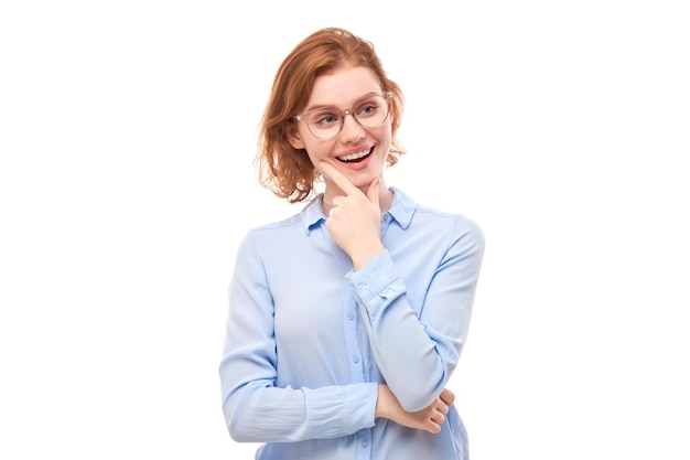 Chica pelirroja inteligente con gafas y camisa de negocios sosteniendo la barbilla piensa que las dudas toman una decisión aislada en el fondo del estudio blanco