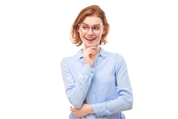 Chica pelirroja inteligente con gafas y camisa de negocios sosteniendo la barbilla piensa que las dudas toman una decisión aislada en el fondo del estudio blanco