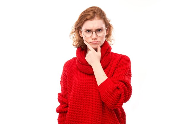 Chica pelirroja inteligente en rojo y gafas sosteniendo la barbilla piensa que las dudas toman una decisión aislada en el fondo del estudio blanco