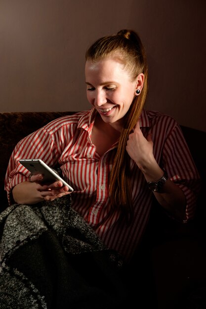 Chica pelirroja jengibre en camisa rayada leyendo el mensaje en el teléfono inteligente, recostado en el sofá y sonriendo