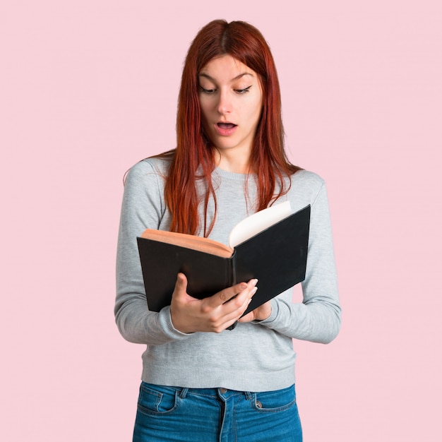 Chica pelirroja joven sosteniendo un libro y sorprendido mientras disfruta de la lectura sobre fondo rosa aislado