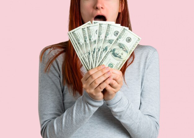 Chica pelirroja joven tomando un montón de dinero en el fondo rosado aislado