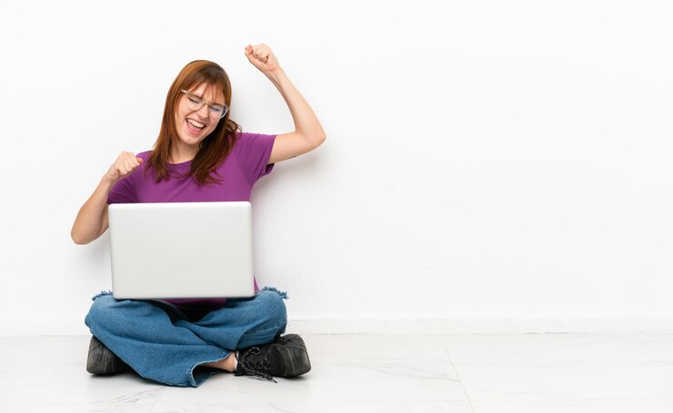 Foto chica pelirroja con una laptop sentada en el suelo celebrando una victoria