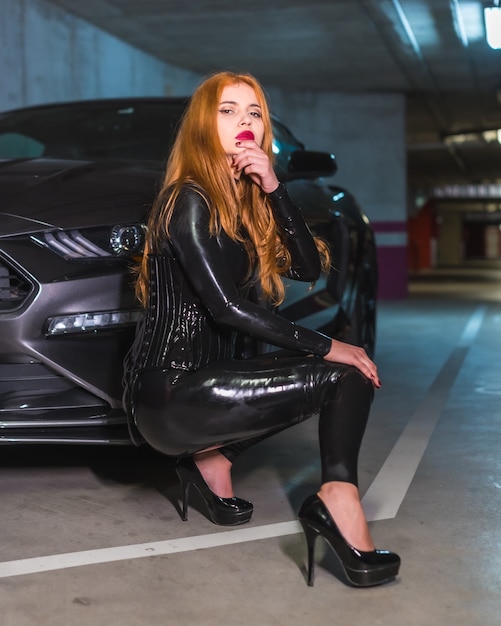 Chica pelirroja en látex, un top negro y pantalones ajustados con tacones grandes, agachada en un auto deportivo en un garaje con mirada seductora