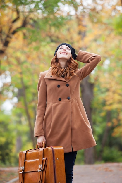 Chica pelirroja con maleta en otoño al aire libre.