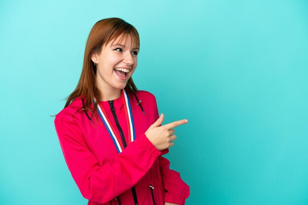 Chica pelirroja con medallas aisladas o fondo azul apuntando con el dedo hacia un lado y presentando un producto