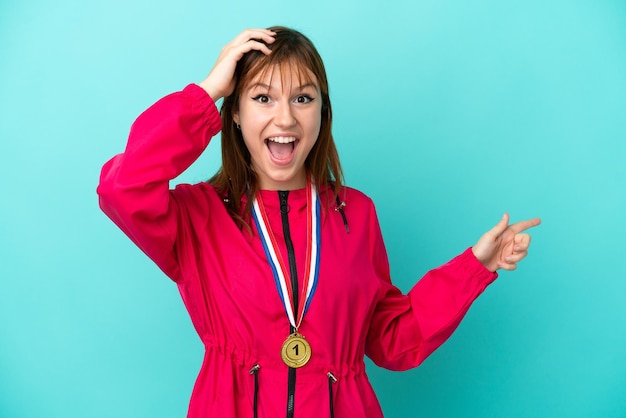 Chica pelirroja con medallas aisladas o fondo azul sorprendida y señalando con el dedo hacia un lado