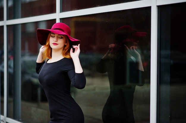 Chica pelirroja de moda con sombrero rosa y vestido negro con maquillaje brillante en una ventana grande