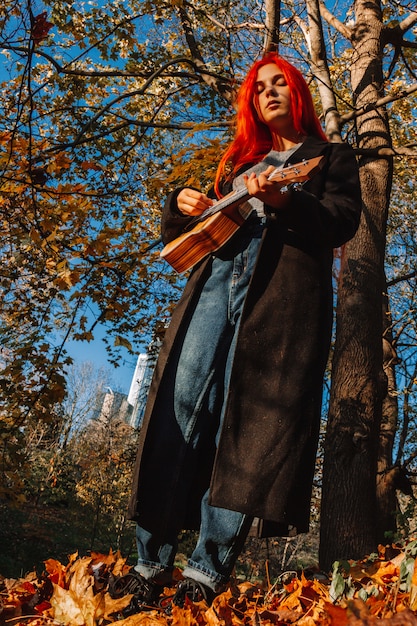 Chica pelirroja con el pelo largo juega en el ukelele en el parque.