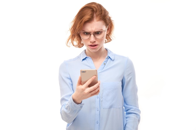 Chica pelirroja de pensamiento pensativo mirando a la pantalla del teléfono inteligente con cara de malestar en gafas y camisa de negocios aislado sobre fondo blanco de estudio