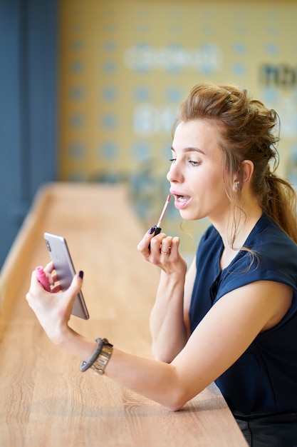 Chica pelirroja poniéndose lápiz labial sola, mirando el reflejo en el teléfono inteligente