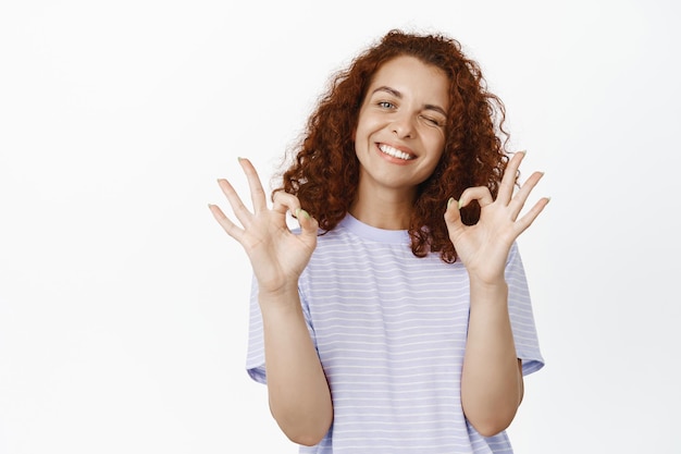 Chica pelirroja positiva guiñando un ojo, sonriendo y mostrando señales de ok, ok, excelente elección, elogiándote, como y de acuerdo, aprueba algo bueno, de pie sobre fondo blanco.