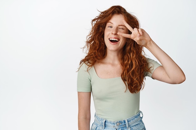 Chica pelirroja positiva muestra un signo de saludo de paz guiñando un ojo y sonriendo a la cámara de pie feliz contra el fondo blanco