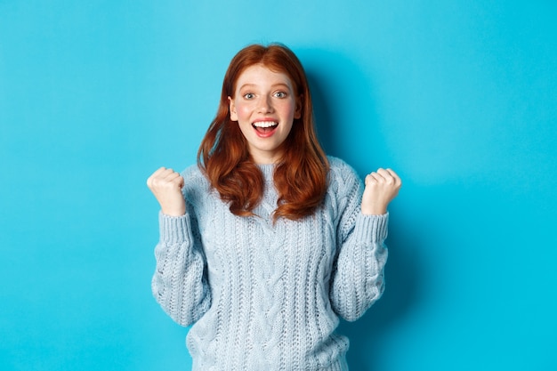 La chica pelirroja satisfecha logra el objetivo y celebra, haciendo un gesto de bomba de puño y sonriendo con regocijo, triunfando de la victoria, de pie contra el fondo azul.