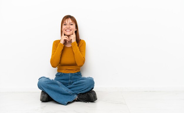 Chica pelirroja sentada en el suelo aislada de fondo blanco sonriendo con una expresión feliz y agradable