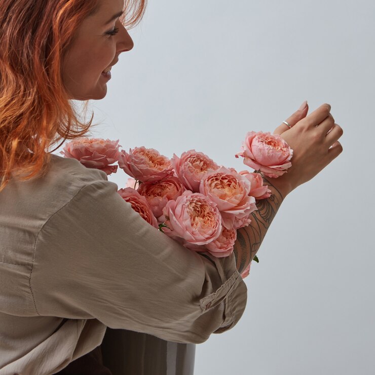 Chica pelirroja sexy con un tatuaje sosteniendo un hermoso ramo de rosas rosadas en un jarrón alrededor de un gris