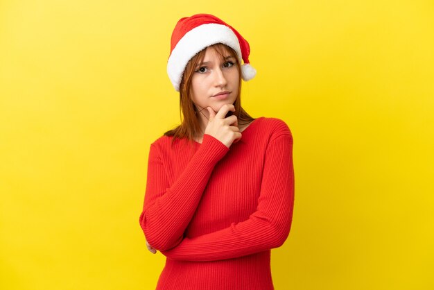 Chica pelirroja con sombrero de navidad aislado sobre fondo amarillo con dudas