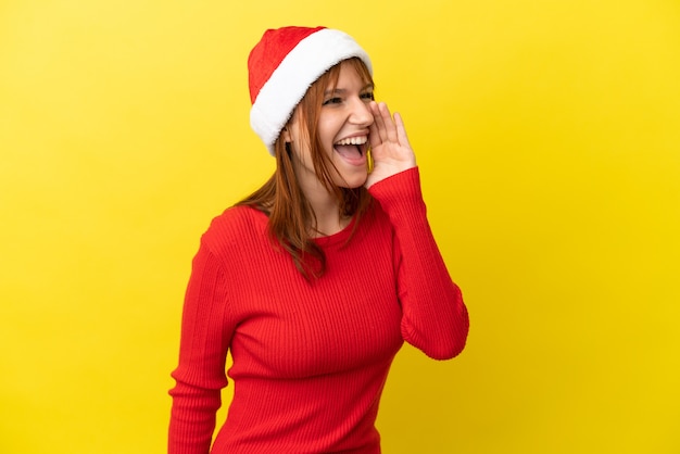 Chica pelirroja con sombrero de navidad aislado sobre fondo amarillo gritando con la boca abierta hacia un lado