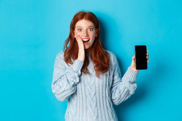 Chica pelirroja sorprendida mirando a cámara, mostrando la pantalla del teléfono inteligente, demostrando la aplicación móvil, de pie sobre fondo azul.