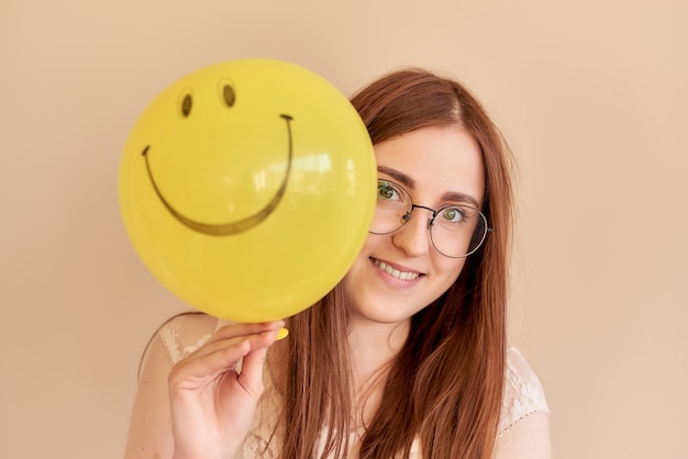 Chica pelirroja sosteniendo un globo amarillo con una sonrisa sobre un fondo beige