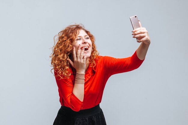 Chica pelirroja toma un selfie en el teléfono sobre un fondo gris claro