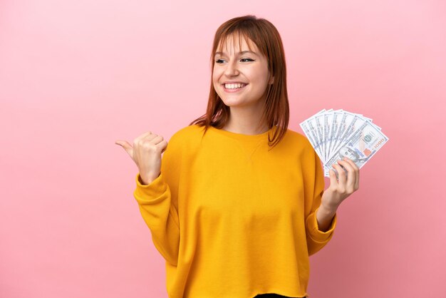 Chica pelirroja tomando mucho dinero aislada en un fondo rosa apuntando hacia un lado para presentar un producto