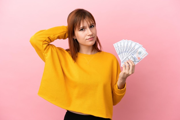 Chica pelirroja tomando mucho dinero aislada de fondo rosa con dudas