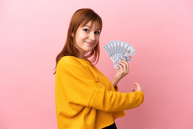 Chica pelirroja tomando mucho dinero aislado sobre fondo rosa apuntando hacia atrás