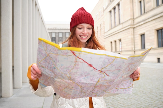 Chica pelirroja turista explora la ciudad mira el mapa de papel para encontrar el camino para los monumentos históricos mujer en
