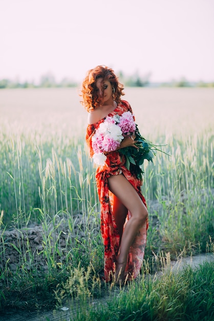 Chica pelirroja en vestido rojo con ramo de peonías bailando alegre en campo de trigo en verano al atardecer. Vertical