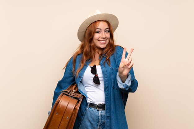 Chica pelirroja viajero con maleta sonriendo y mostrando el signo de la victoria