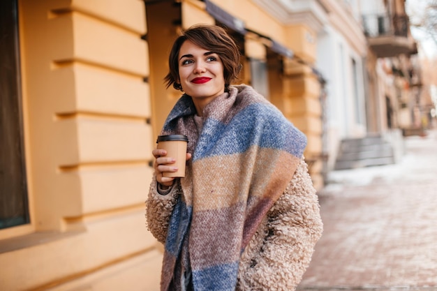 Chica de pelo corto inspirada bebiendo café en la calle Disparo al aire libre de una mujer morena con abrigo