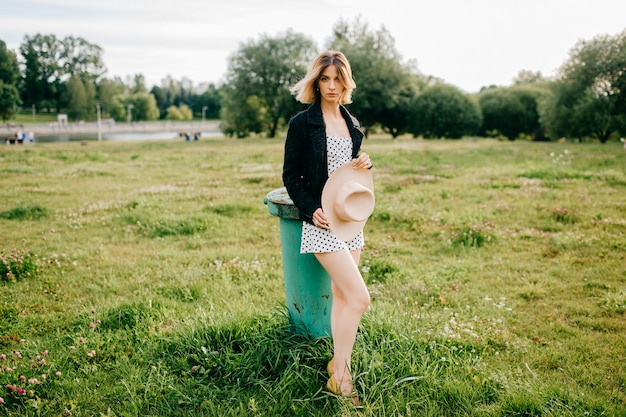 Chica de pelo corto rubia con estilo elegante posando con sombrero en el campo