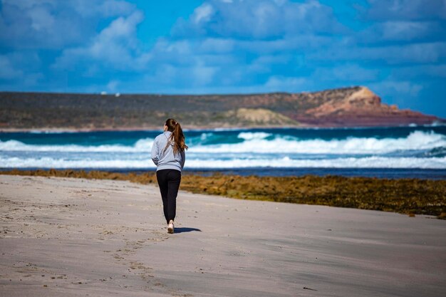 una chica de pelo largo camina por la playa con enormes acantilados al fondo en el parque nacional de kalbarri