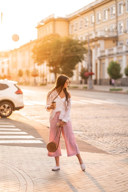 Chica de pelo largo con gafas con café de teléfono y bolso camina por la ciudad al atardecer