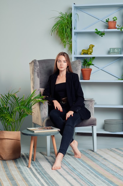 Chica con el pelo largo en jeans y chaqueta posando en el sillón en la sala de estar