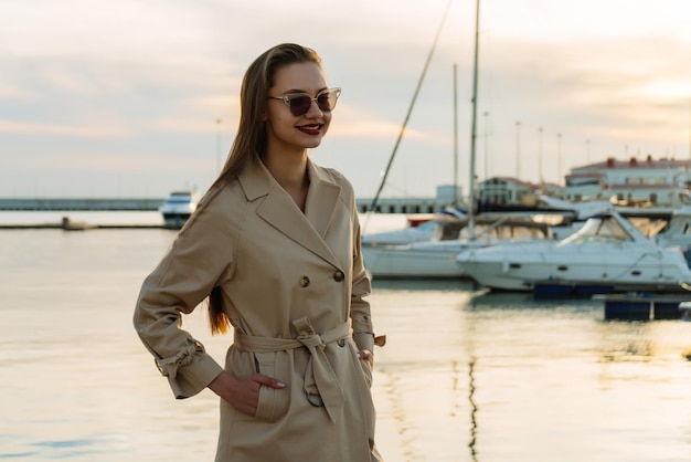 Chica de pelo largo de moda con un elegante abrigo beige y gafas negras posando en el puerto marítimo