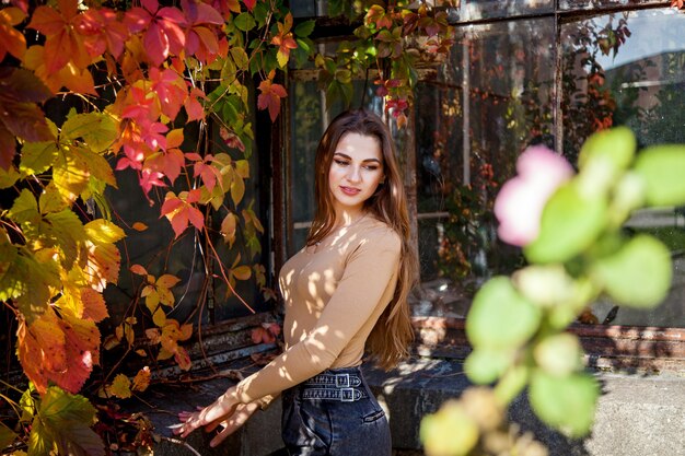 Una chica con el pelo largo en un mono y jeans posando en un parque de otoño con el telón de fondo de una pared cubierta de uvas silvestres. Chicas en el parque en el fondo de uvas silvestres