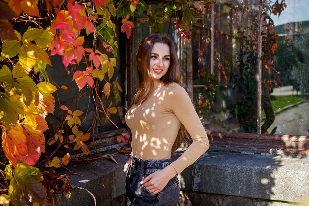 Una chica con el pelo largo en un mono y jeans posando en un parque de otoño con el telón de fondo de