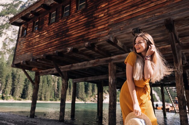 Chica con el pelo largo posando cerca de un edificio de madera junto al lago de montaña. mujer joven en vestido y navegante.