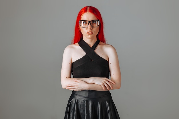 Chica con el pelo largo rojo con gafas y una falda de cuero posando sobre un fondo gris