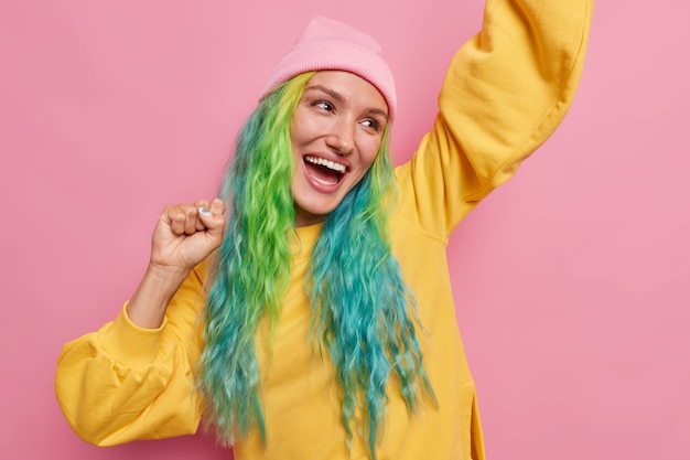 chica con el pelo largo teñido hace bailar al campeón levanta las manos mira felizmente lejos se siente como el ganador usa sombrero y jersey amarillo aislado en rosa