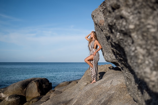 Chica de pelo largo con un vestido de rayas descansando sobre las rocas cerca del mar. Vacaciones de verano. Phuket, Tailandia