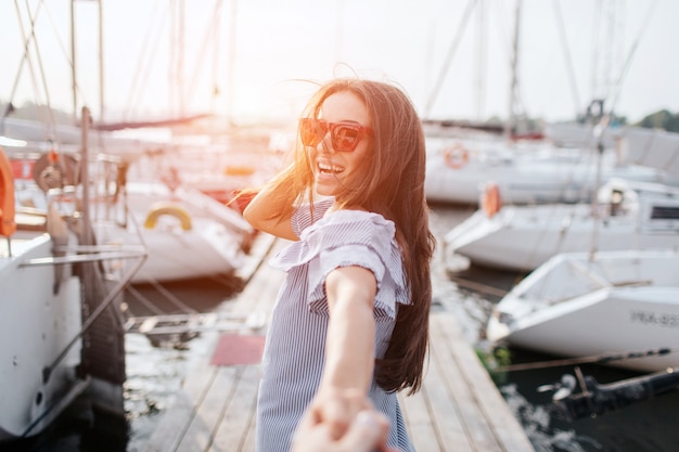 Chica de pelo oscuro se encuentra en el muelle y sonriendo. Su cabello está ondeando. Ella usa gafas de sol y toma la mano. La morena lleva un vestido a rayas. Ella luce feliz. Sígueme.