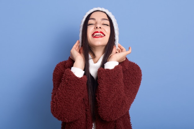 Chica de pelo oscuro se pone el sombrero de invierno sobre espacio azul aislado riendo