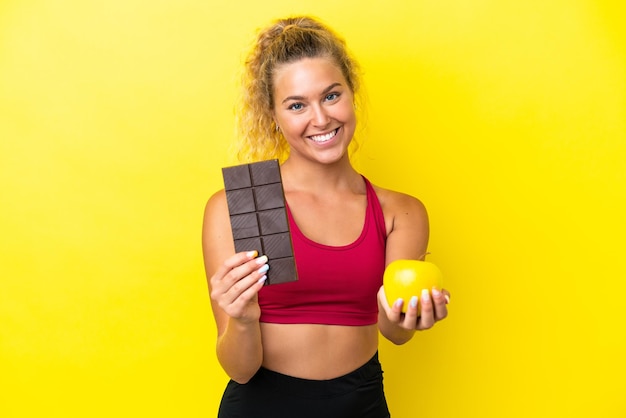 Chica con el pelo rizado aislada de fondo amarillo tomando una tableta de chocolate en una mano y una manzana en la otra