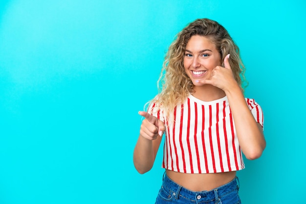 Chica con el pelo rizado aislado sobre fondo azul haciendo gesto de teléfono y apuntando hacia el frente